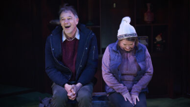 A man and woman sit on a bench. The man, wearing a jacket, smiles and laughs, whilst the woman, in a knitted hat and waistcoat, looks down and smiles shyly. They appear to be sharing a light-hearted moment together.