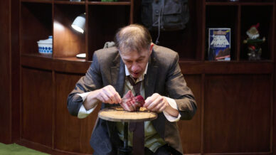 A man in a suit sits on a stool, intensely examining and manipulating a piece of raw meat on a small round table. Shelves with various objects are visible in the background, including a Star Wars book.