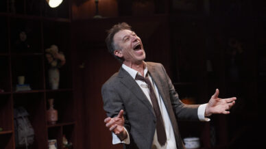 A man in a grey suit and loosened tie stands indoors with his mouth open wide and eyes closed, appearing to sing or shout passionately. He gestures with both hands. Shelves and dim lighting are visible in the background.