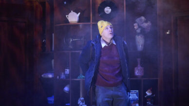 A person wearing a yellow beanie, maroon jumper, and blue jacket stands in front of a wooden shelf filled with various objects, including vases, a teapot, and figurines, surrounded by a misty atmosphere.