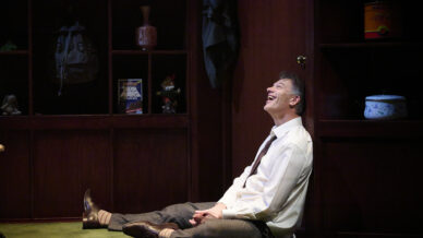 A man in a white shirt and tie sits on the floor, leaning against a wooden cupboard, laughing. Behind him are shelves with various objects, including books, a jar, and a rucksack.