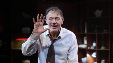 A man in a white shirt and loose dark tie stands indoors, holding his hand up with fingers spread, as if waving or signalling. He is smiling slightly, and shelves with various objects are visible in the dim background.