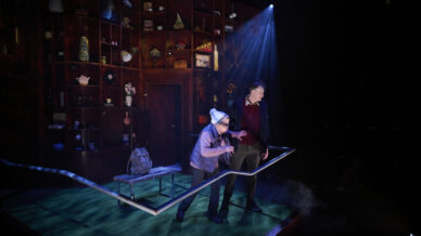Two actors perform on a stage with dramatic lighting; one leans on a railing, wearing a white hat, whilst the other stands beside them. The backdrop is filled with shelves holding various objects and decorations.
