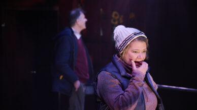 A woman in a knitted hat and waistcoat stands in the foreground, looking concerned with her hand near her mouth. A man in a jacket is blurred in the background, standing and looking off to the side. The lighting is dramatic.