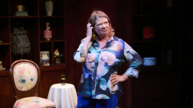 A woman in a colourful blouse stands with one hand on her hip and the other touching her head, looking puzzled. She is on stage in a cosy room with wooden shelves, a floral chair, and a small round table.