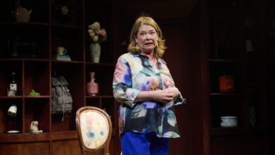 A woman stands on stage in front of wooden shelves filled with various objects. She wears a colourful blouse and blue trousers, with a patterned chair beside her, looking expressive as if speaking or performing.