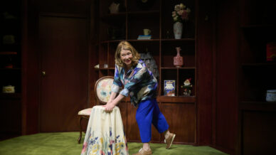 A woman with shoulder-length hair, wearing a blue blouse and trousers, leans forward with one hand on a chair covered by a floral cloth in a room with wooden shelves and decorative items.