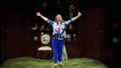 A woman stands on a green carpet with her arms raised and smiling, in front of wooden shelves filled with decorative items and a chair. She wears a floral shirt, blue trousers, and brown shoes.