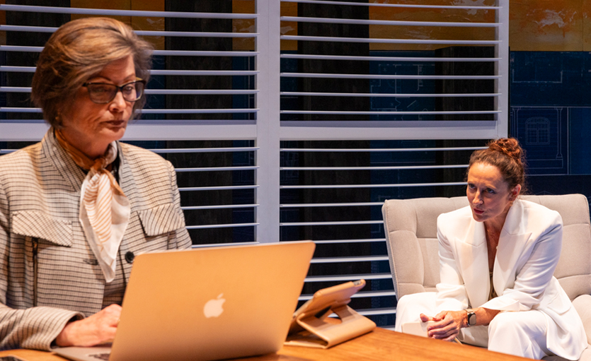 Two women in business attire are in an office. One sits at a desk working on a laptop, looking serious. The other, dressed in white, sits on a chair in the background, appearing to listen or speak thoughtfully.