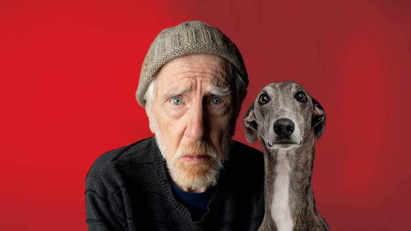 An elderly man wearing a knitted hat and jumper sits next to a greyhound against a bright red background, both looking directly at the camera with serious expressions.