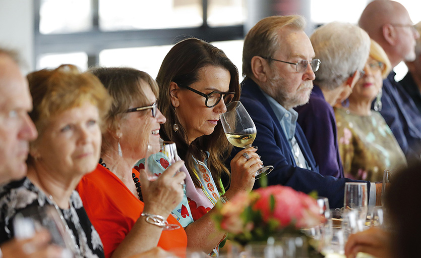 A group of people sit at a dining table, enjoying drinks and food. A woman in the centre smells a glass of white wine. The atmosphere appears lively and social, with flowers on the table.