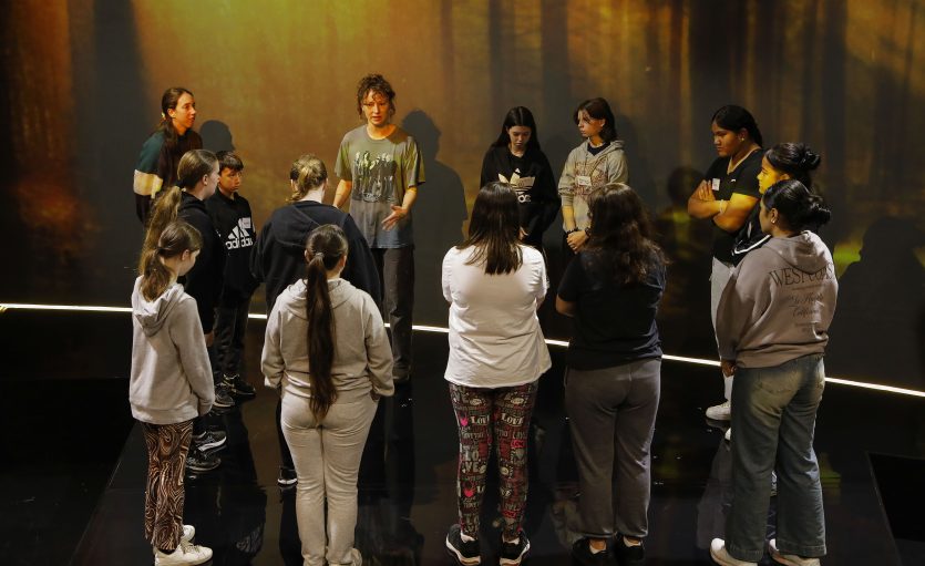 Smiling older man with grey hair and beard wearing a blue blazer over a white T-shirt featuring a black-and-white portrait of another elderly man, standing in front of a solid red background.