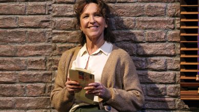 A smiling woman leans against a brick wall, while holding a ripped open envelope. The envelope address has been hand-written and is covered in a significant amount of stamps.