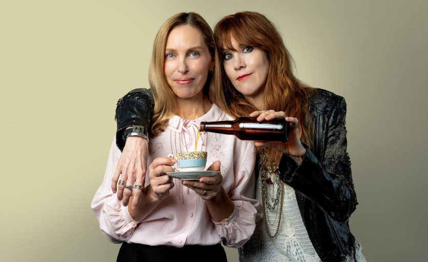 Womenin pink blouse holding a teacup, second woman in gungy leather jacket leaning on her pouring beer from a bottle into the teacup.