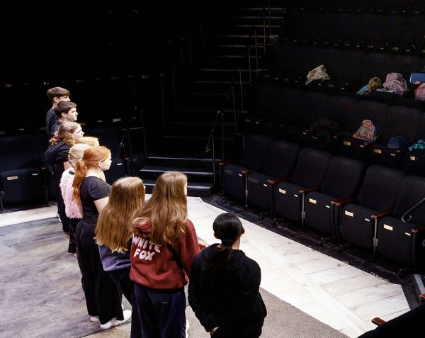 A group of students with their back to the camera looking out at an empty auditorium. A teacher stands in the seating bank talking to them.