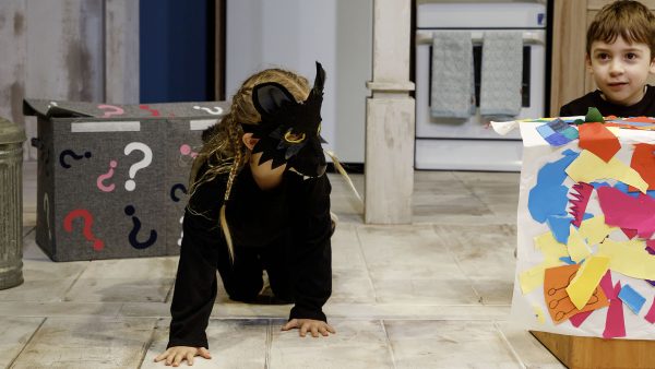 Young girl wearing wolf mask and young boy sitting behind box covered in colourful collage.