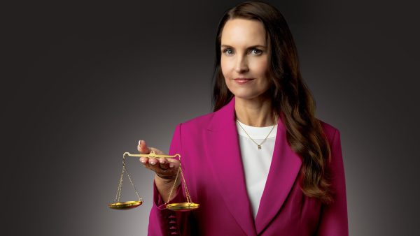 A woman in a white t-shirt and a pink blazer looks at the camera and holds a gold scale of justice in her hands.
